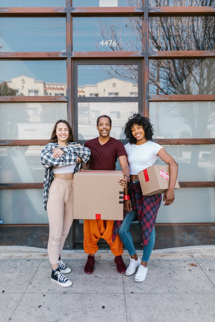 Three friends smiling while holding cardboard boxes, enjoying the experience of moving into a new home.